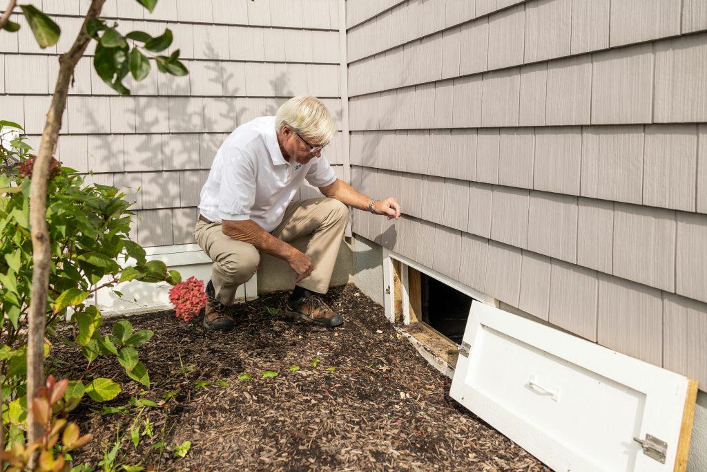 man peering through the opening of a crawl space beneath the exterior of a house